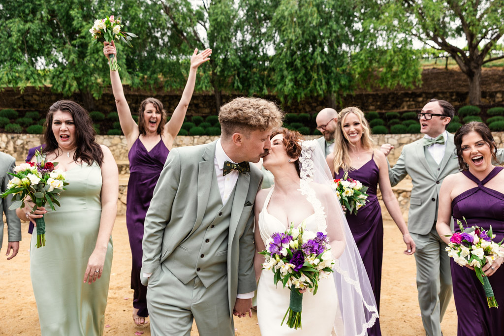 Bride and groom kiss outdoors at a California vineyard wedding, surrounded by a joyful wedding party in purple and green dresses and suits