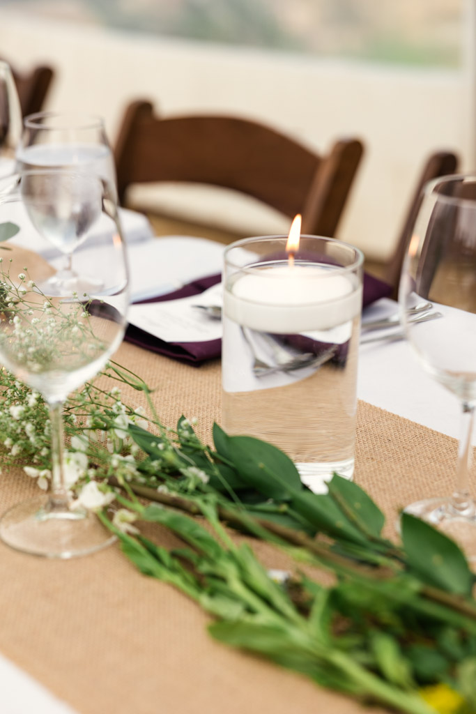 A lit floating candle in a glass sits on a guest table with greenery, wine glasses, and a burlap runner for wedding reception at Terra Mia Vineyard in Paso Robles, California