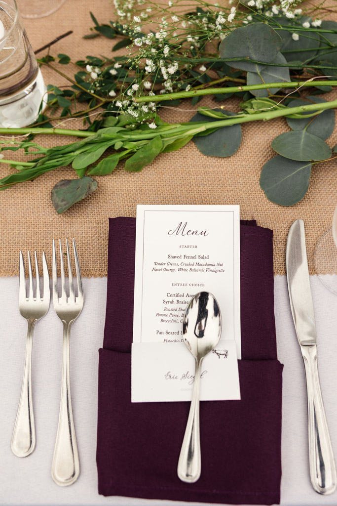 Guest table place setting with a menu on a purple napkin, silver cutlery, and greenery centerpiece for wedding reception at Terra Mia Vineyard