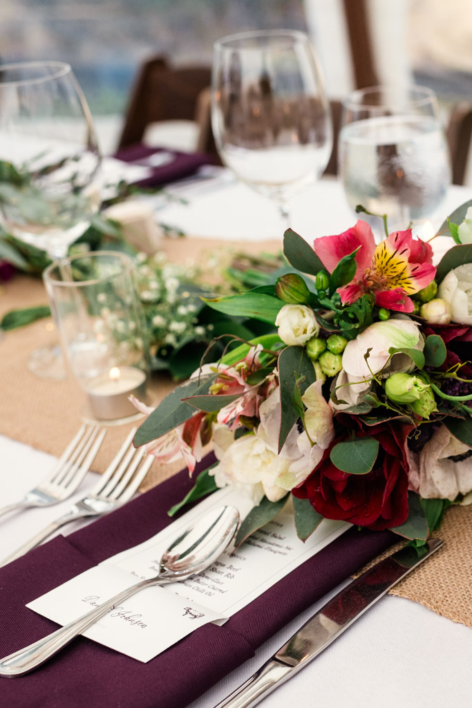 Detail photo of guest table decor at a Terra Mia Vineyard wedding reception, featuring wine glasses, cutlery, a floral centerpiece, and a purple napkin