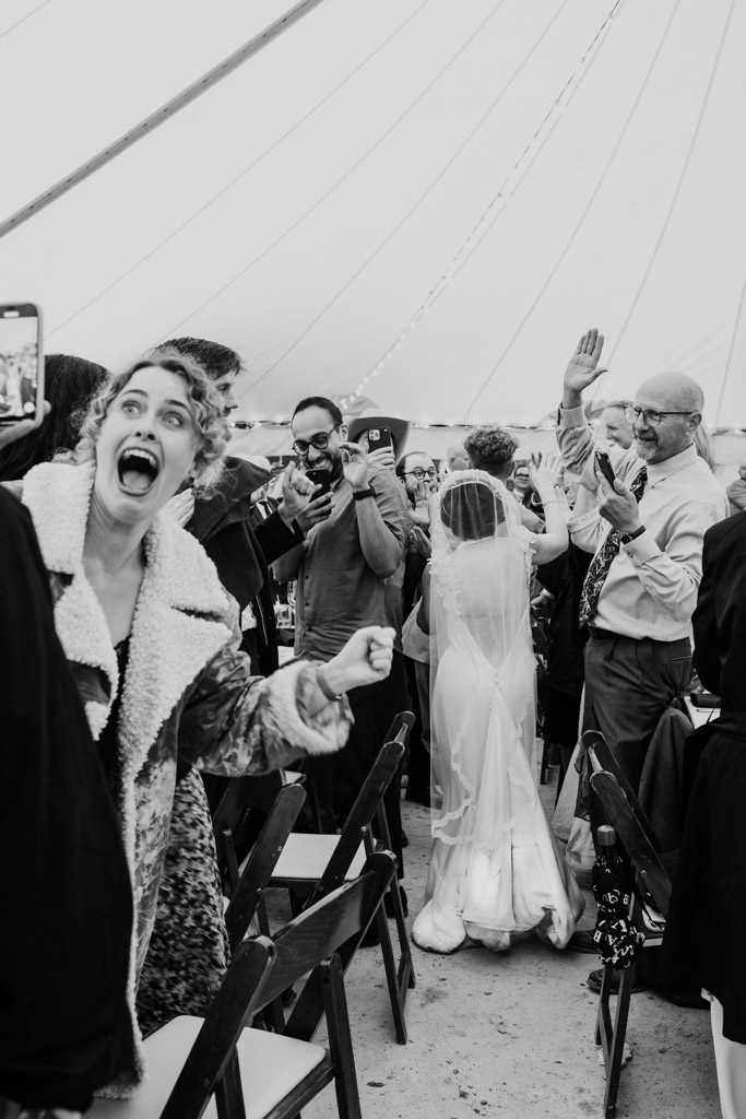 Black and white photo of joyful woman cheering as bride walks among the guest tables high-fiving guests during Terra Mia Vineyard wedding reception