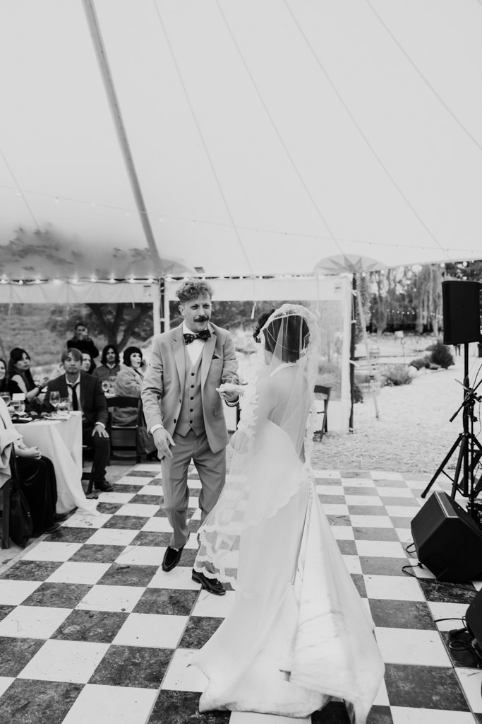 Black and white photo of bride and groom dance together at their Terra Mia Vineyard wedding reception under a tent, surrounded by seated guests
