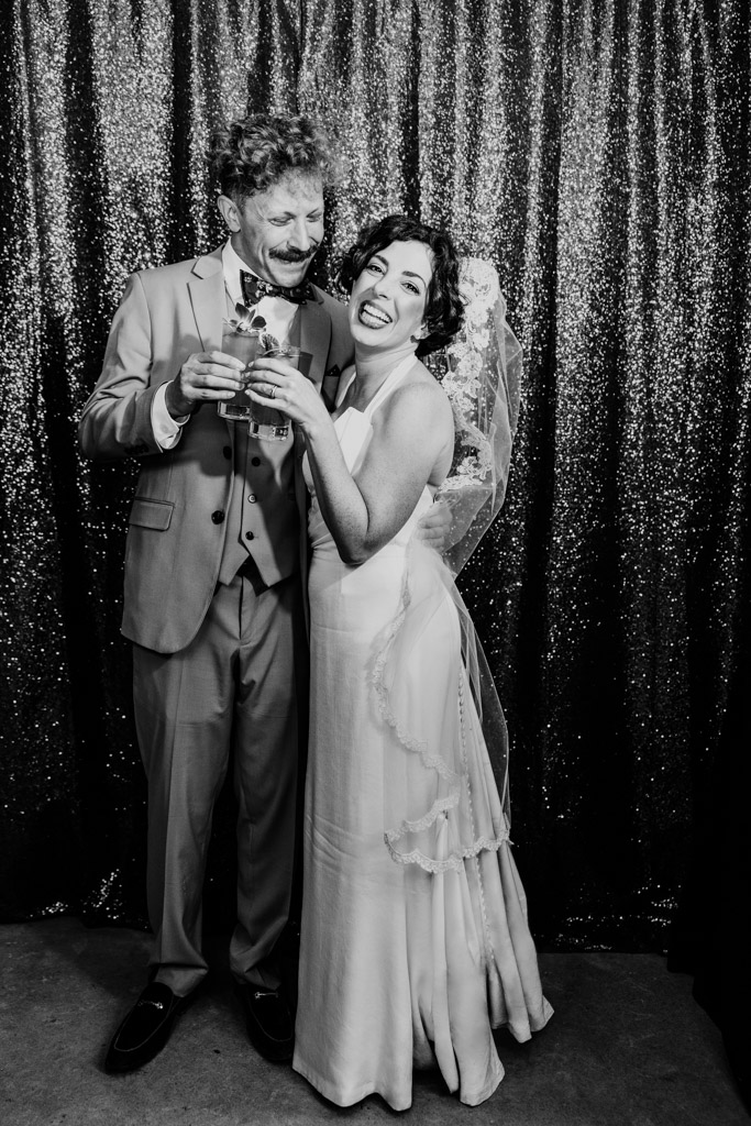 Black and white photo of happy bride and groom laughing together, holding drinks in front of a sparkly curtain at their wedding reception at Terra Mia Vineyard