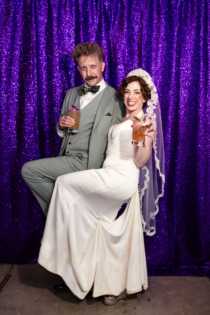 Portrait of bride and groom smiling with drinks, posing playfully in front of a sparkly purple curtain at their Terra Mia Vineyard wedding celebration