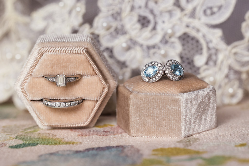 Velvet ring boxes with diamond rings and blue gemstone earrings rest on a floral surface with bride's veil in the background for wedding celebration at Terra Mia Vineyard in Paso Robles, California