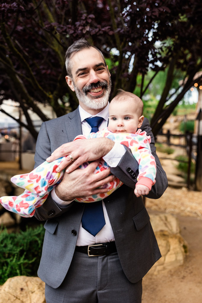 Smiling man in a suit holds a baby in a heart-patterned outfit outdoors at Terra Mia Vineyard wedding reception