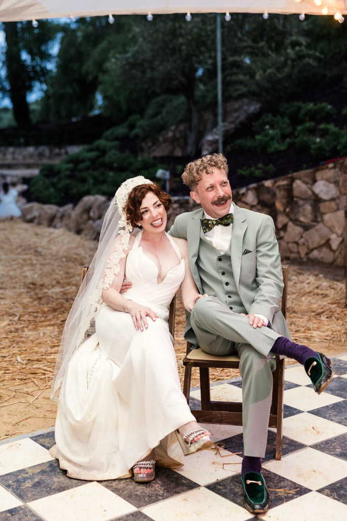 Happy bride and groom sit together on chairs outdoors on a black-and-white checkered floor during their Terra Mia Vineyard wedding reception