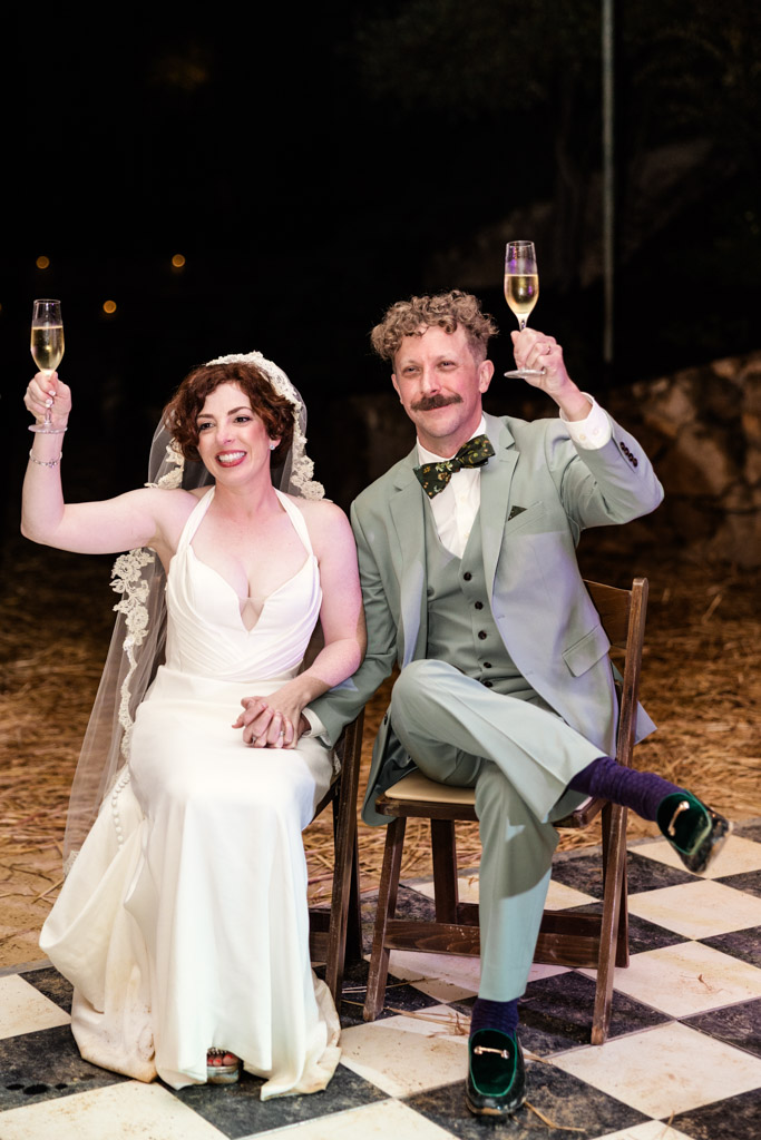 Bride and groom sitting on chairs, smile and raise champagne glasses in a celebratory toast at their Terra Mia Vineyard wedding reception