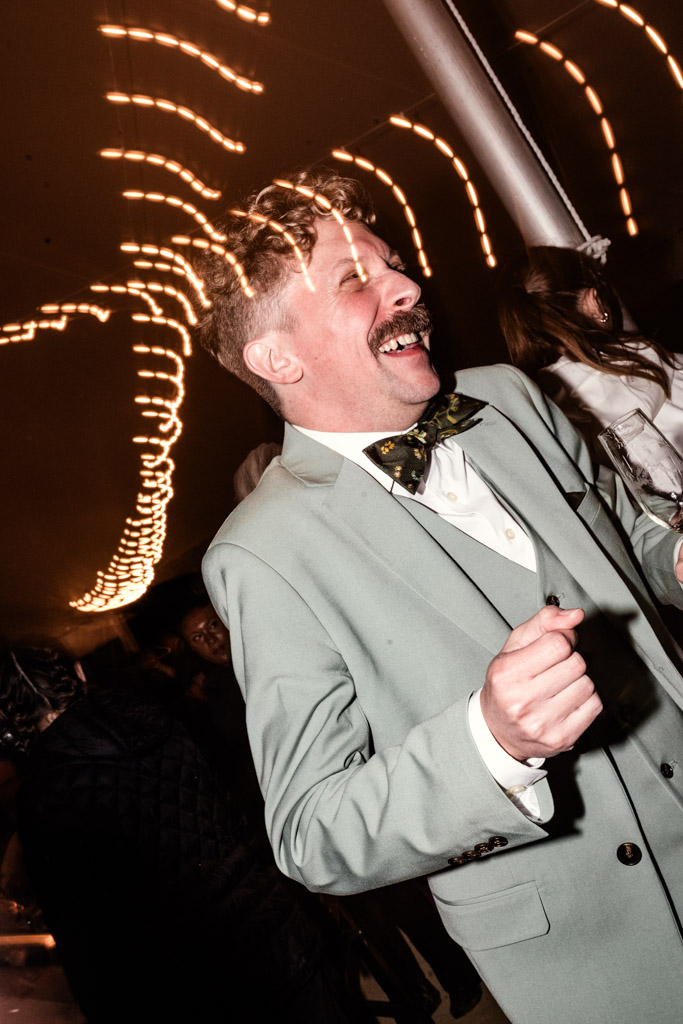 Groom in a light green suit laughs and dances under string lights at his Terra Mia Vineyard wedding reception