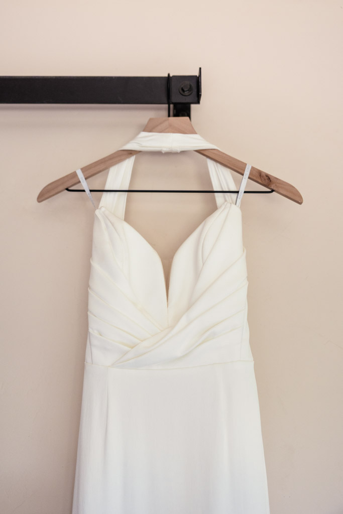 Bride's sleeveless white wedding dress with a deep neckline hangs on a wooden hanger against a beige wall fir wedding at Terra Mia Vineyard in Paso Robles, California