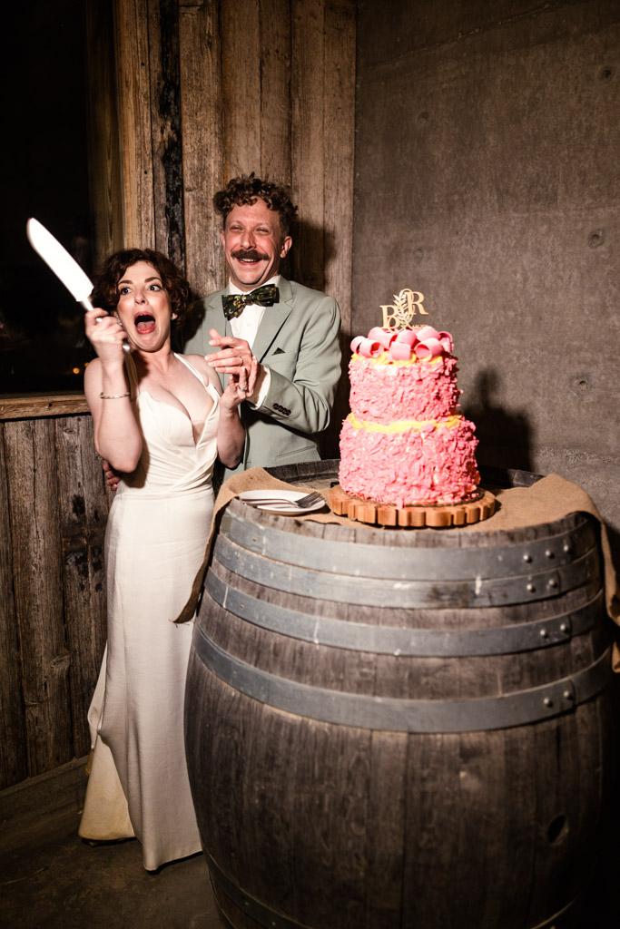 Happy newlyweds in wedding attire prepare to cut a pink tiered cake on a barrel table during their Terra Mia Vineyard wedding reception