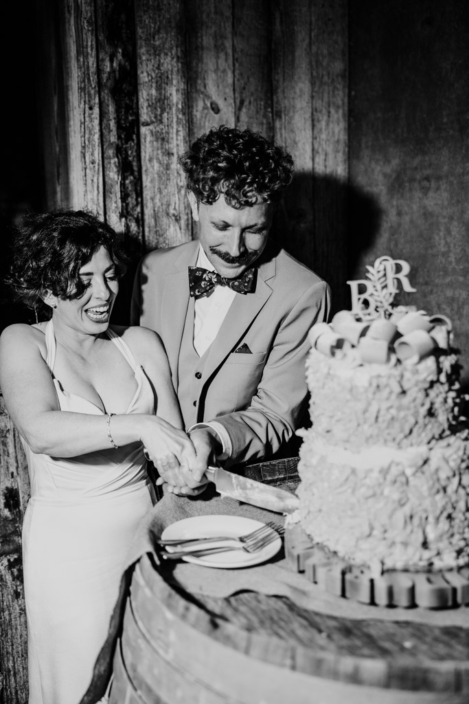 Black and white photo of newlywed couple in formal attire smiling as they cut a wedding cake together on a rustic wooden barrel at their Terra Mia Vineyard wedding reception