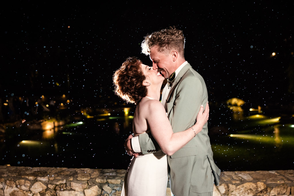 Newlywed couple embraces and smiles at night under falling rain, with lights glowing softly in the background during their wedding reception at Terra Mia Vineyard