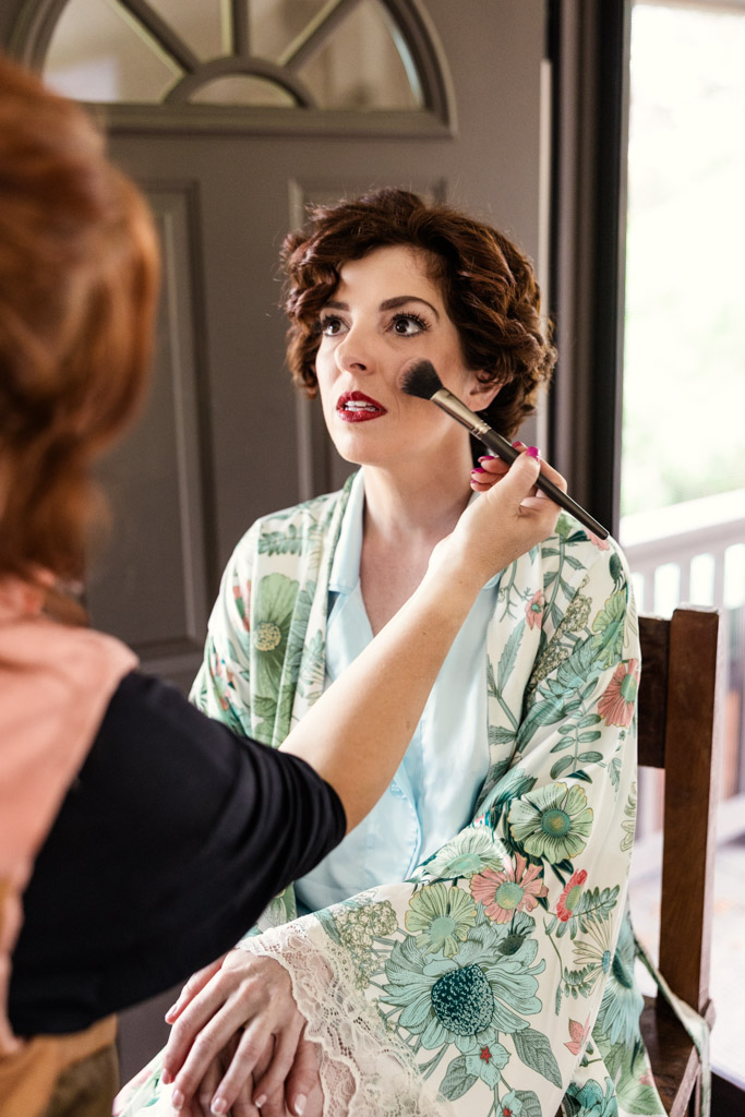 Bride in a floral robe gets her makeup done with a brush on her cheek before her Terra Mia Vineyard wedding celebration