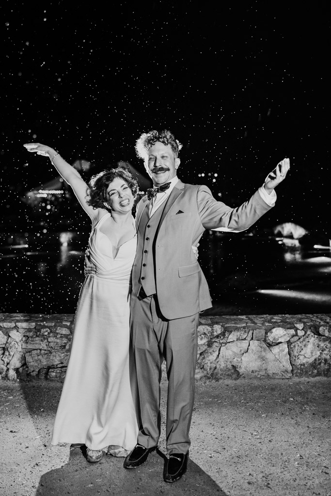 Black and white photo of happy newlyweds in formal attire arms raised in the rain at night during their wedding celebration at Terra Mia Vineyard in Paso Robles, California