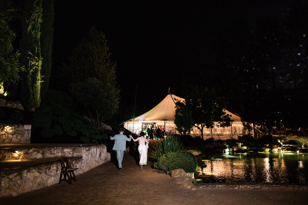 Newlywed couple walks hand in hand toward a glowing tent at night beside a pond during their wedding reception at Terra Mia Vineyard