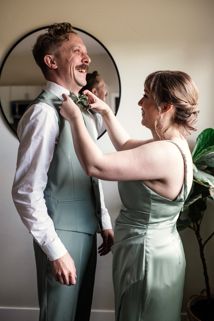 Maid of Honot adjusts groom's bowtie as they both smile, wearing matching sage green formal outfits for wedding celebration at Terra Mia Vineyard