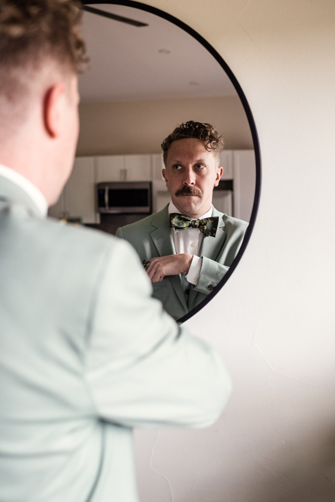 Groom in a light green suit with a bow tie looks at his reflection in a round mirror on a beige wall before his wedding celebration at Terra Mia Vineyard