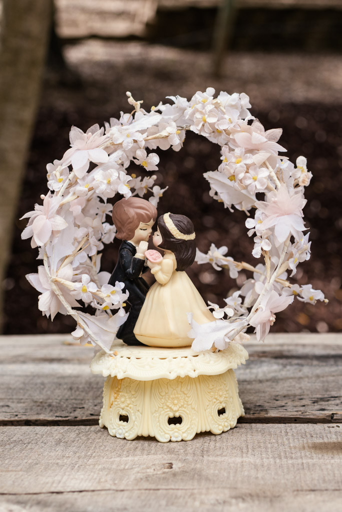 A wedding cake topper shows a bride and groom figurine under an arch of delicate white flowers on a wooden table for Terra Mia Vineyard wedding celebration