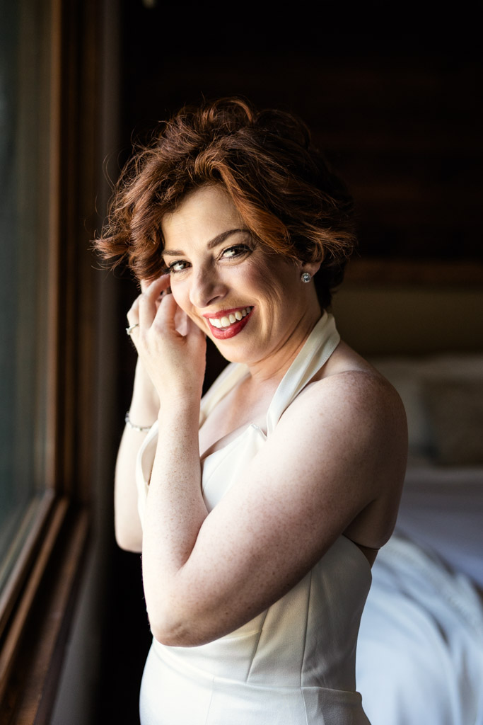 Happy bride in a white dress stands by a window while putting on her earring, with natural light on her face before her Terra Mia Vineyard wedding