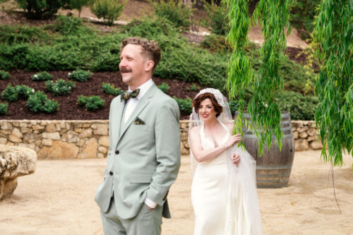 A bride in a white dress smiles as she approaches a groom in a light suit for a first look at their California vineyard wedding.