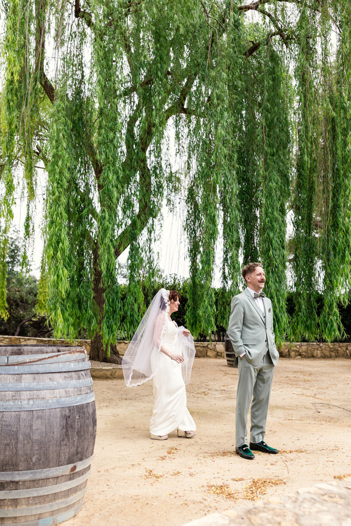 Bride walks toward groom beneath a large tree during their first look at Terra Mia Vineyard
