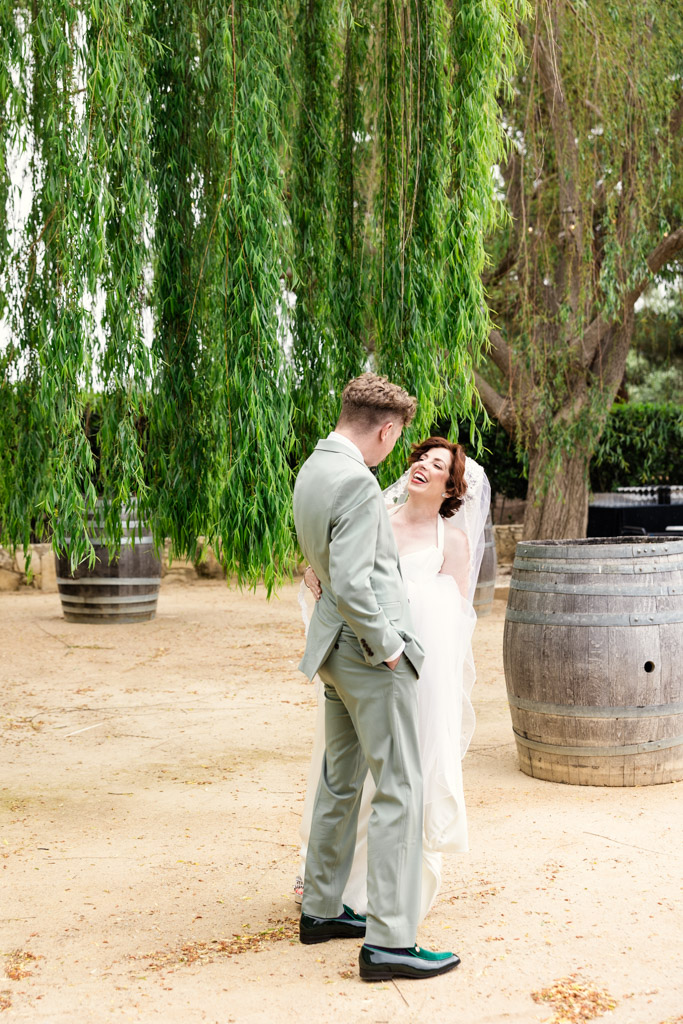 A bride and groom laugh together beneath a large tree during their first look at Terra Mia Vineyard