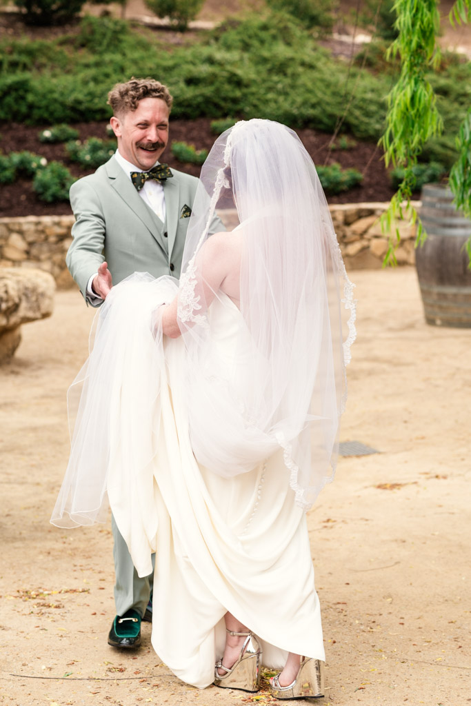 Groom in a light suit smiles as he greets his bride in a white dress and veil during their first look at Terra Mia Vineyard