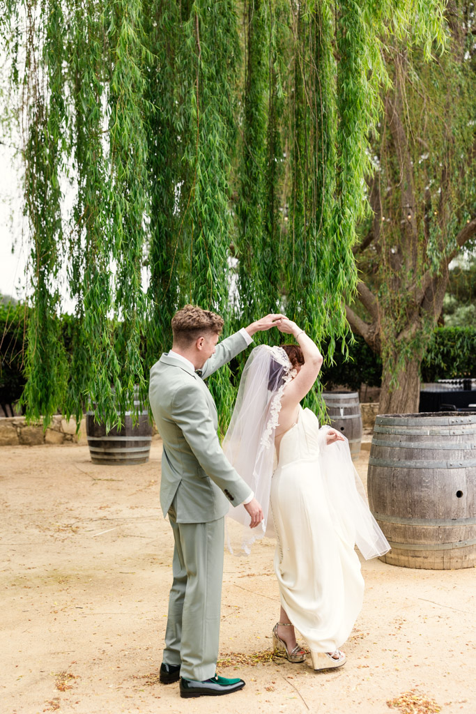 Groom in a light green suit twirls his bride in a white dress and veil under hanging willow branches at Terra Mia Vineyard in Paso Robles, California