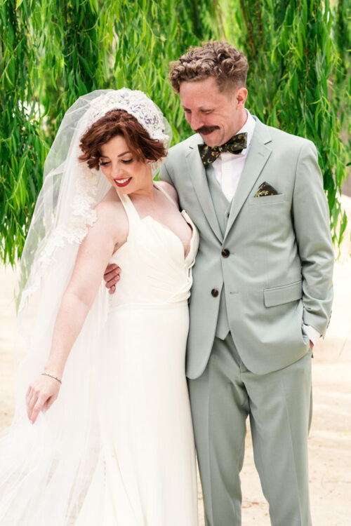 Bride in white dress and veil stands smiling with groom in light grey suit under green leafy branches.