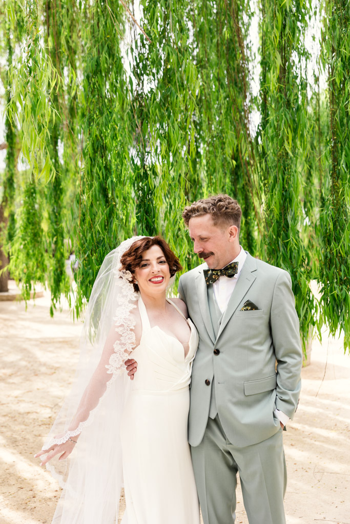 Bride and groom smiling under green willow trees at Terra Mia Vineyard, bride in white dress, groom in light green suit with bow tie