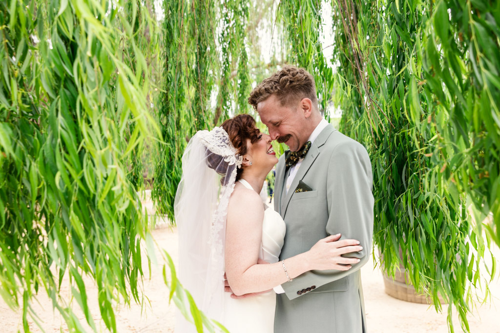 A bride and groom embrace and smile under hanging willow branches on their wedding day at Terra Mia Vineyard