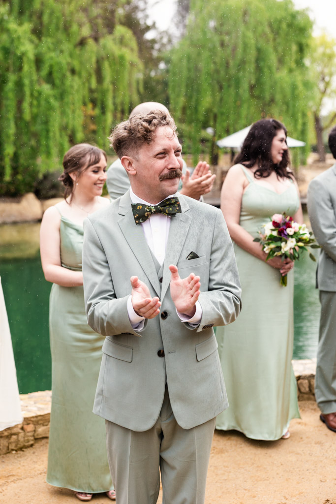 Groom in a pale green suit claps, standing with bridesmaids in matching dresses by a pond during wedding ceremony at Terra Mia Vineyard
