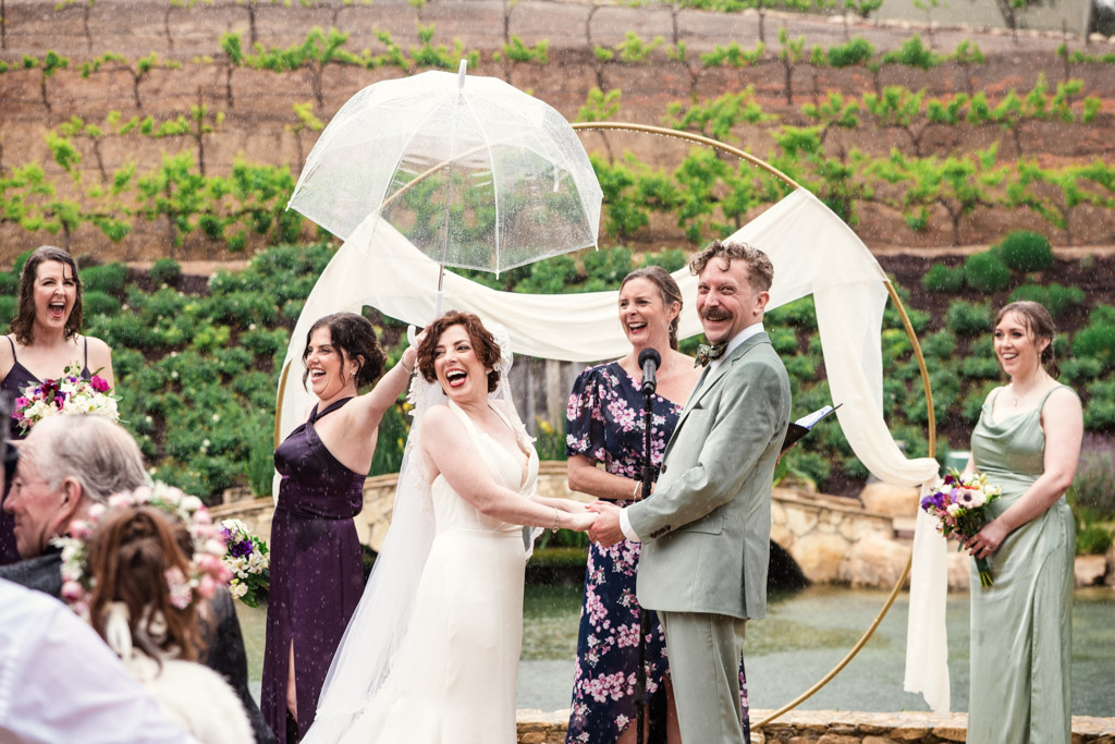 Bride and groom laughing in the rain at their Terra Mia Vineyard wedding, with a bridesmaid holding an umbrella