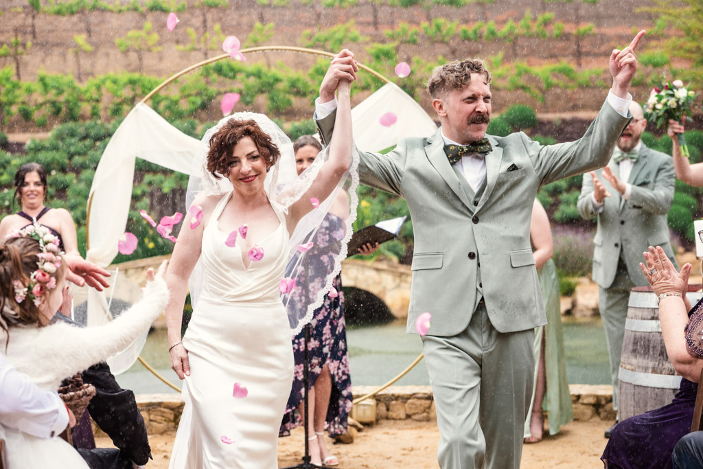 Bride and groom walk down the aisle after their wedding ceremony at Terra Mia Vineyard as guests toss pink flower petals