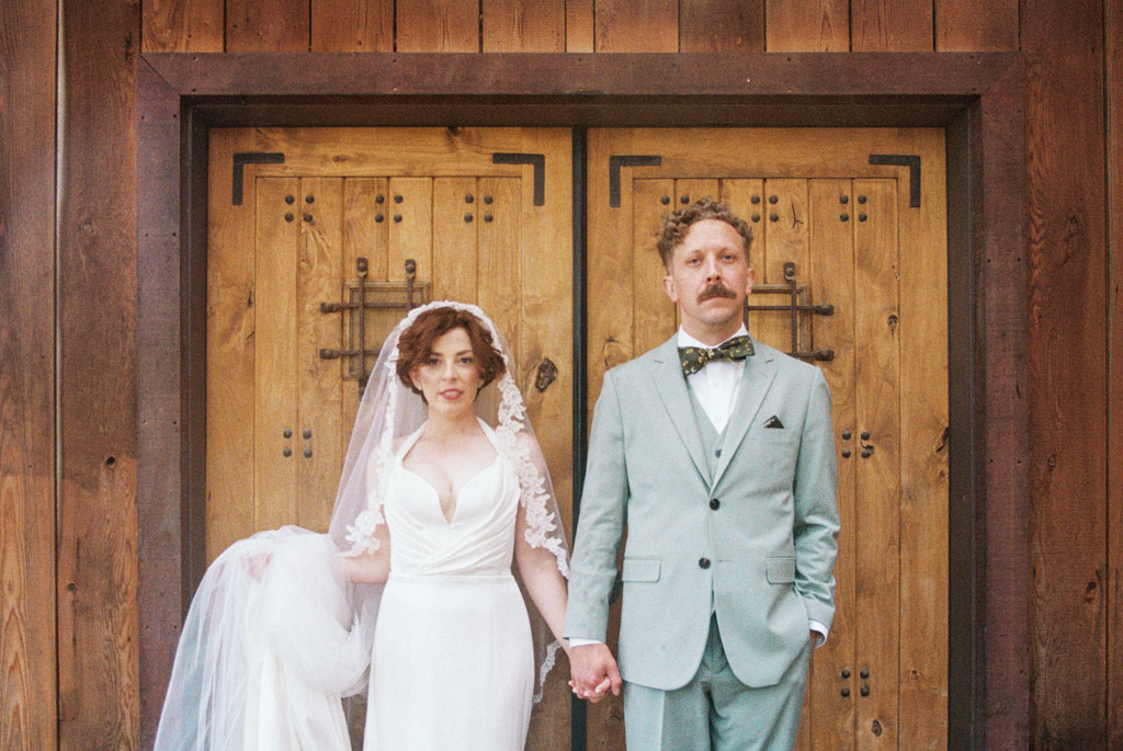 Film portrait of bride in a white dress and groom in a light green suit holding hands in front of wooden doors at Terra Mia Vineyard