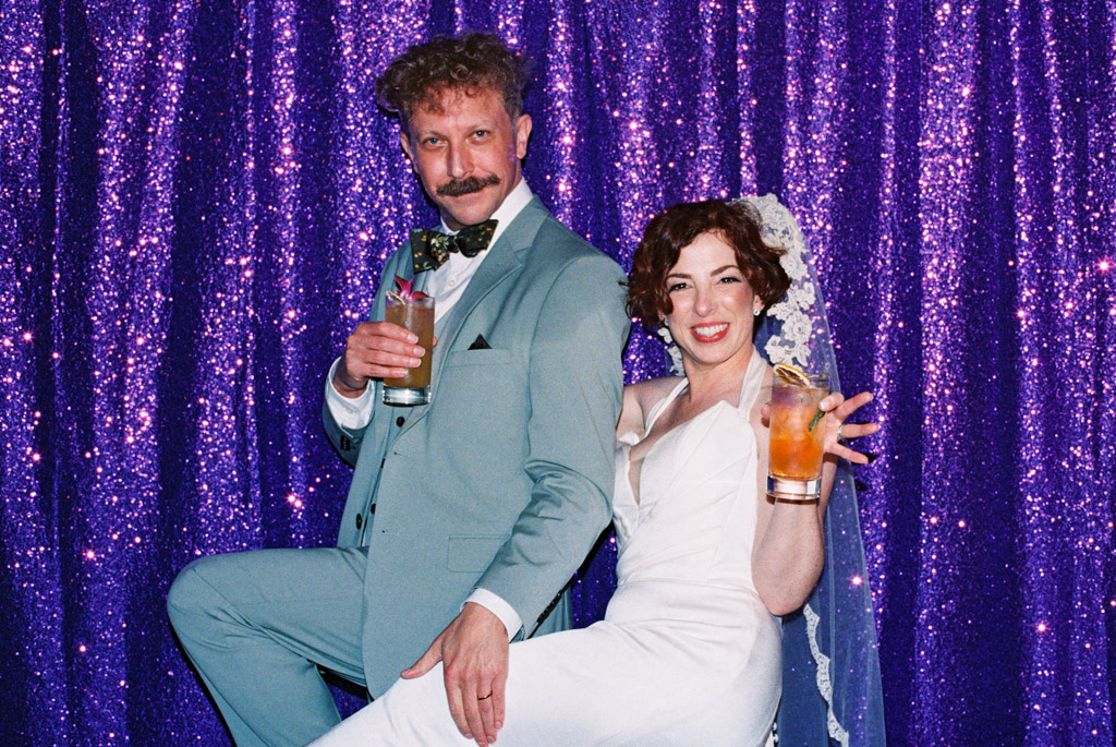 Film photo of smiling newlywed couple in formal attire posing with drinks in front of a sparkly purple sequin backdrop during their wedding reception at Terra Mia Vineyard