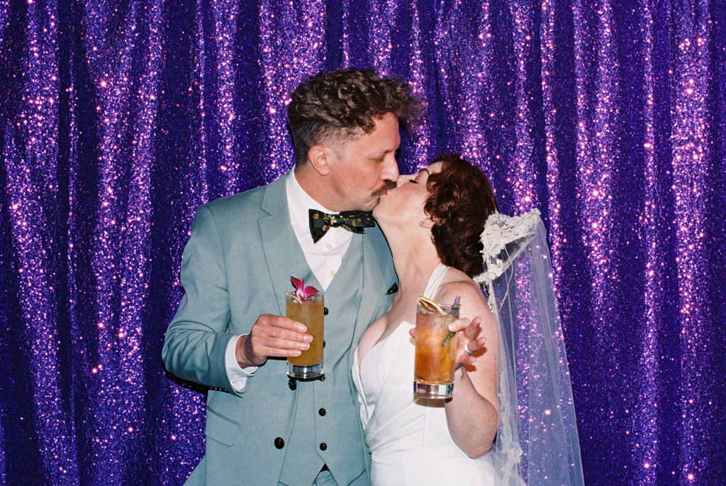 Film photo of bride and groom kissing while holding cocktails in front of a sparkling purple sequin backdrop at their Terra Mia wedding reception