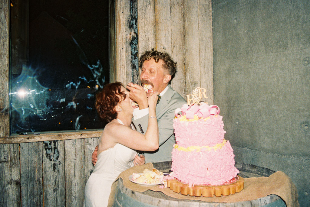 Film photo of bride and groom laughing while feeding each other pink wedding cake during their wedding reception at Terra Mia Vineyard