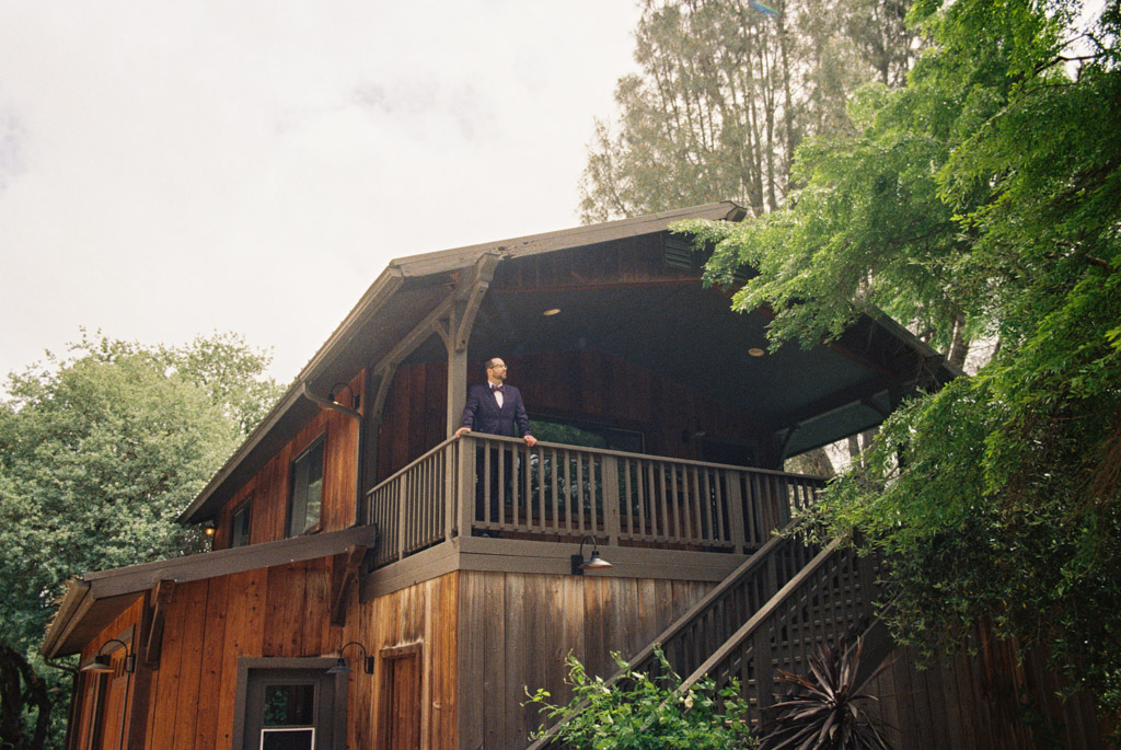 Film photo of groomsman in a purple suit stands on the balcony of a wooden house surrounded by trees at Terra Mia Vineyard in Paso Robles, California