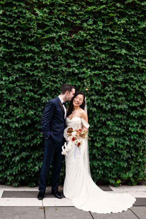 A bride and groom stand smiling in front of a lush green ivy wall on their wedding day.
