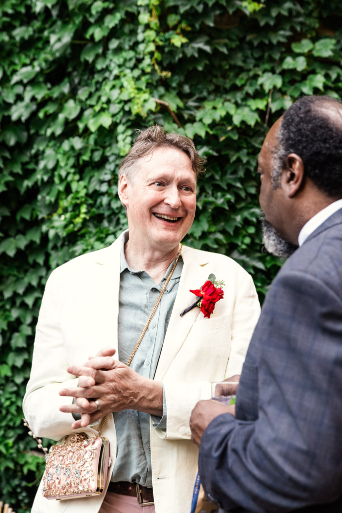 Groom wearing a white jacket with a red flower talks with a guest in front of an ivy-covered wall during his wedding reception at Firehouse Chicago
