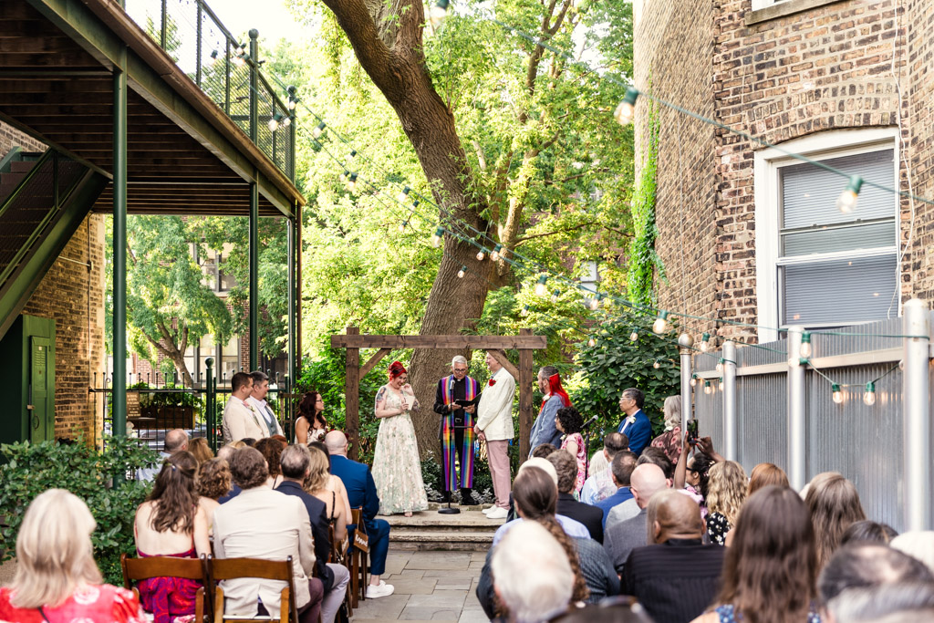 Outdoor wedding ceremony at Firehouse Chicago, with guests seated as the couple and officiant stand beneath a wooden arbor surrounded by trees in the background
