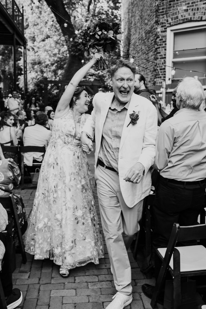 Black and white photo of happy newlywed couple walking hand in hand down the aisle at their outdoor Firehouse Chicago wedding ceremony , surrounded by cheering guests