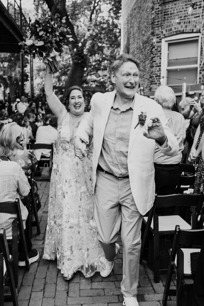 Black and white phot oof newlywed couple walking down the aisle, smiling and celebrating at their outdoor Firehouse Chicago wedding ceremony