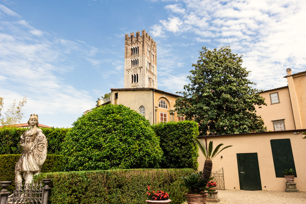 A stone tower rises behind green hedges, a statue, and a tan building under a partly cloudy sky in Lucca, Italy