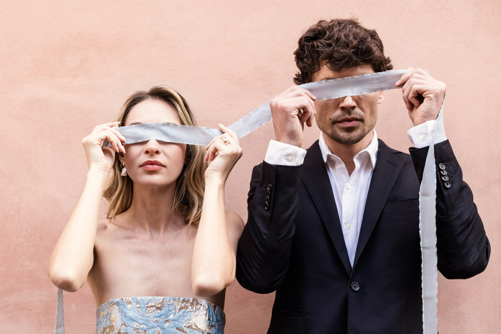 A woman and man hold a white ribbon over their eyes against a pale pink wall in Lucca, Italy