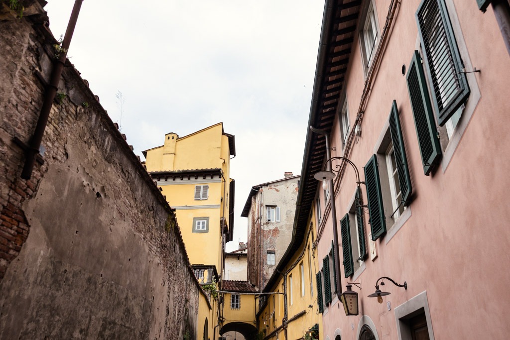 Narrow street in Lucca, Italy lined with pastel buildings, green shutters, and street lamps under a cloudy sky