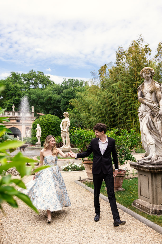 A couple dressed formally smiles and holds hands while walking in Orto Botanico Comunale di Lucca with statues and greenery