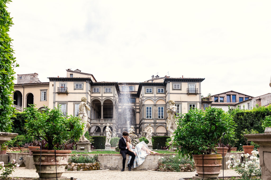 A couple sits together in a Lucca, Italy garden courtyard with a fountain and a grand villa in the background